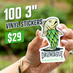 Hand holding a die-cut sticker of a stylized trout with green scale-like patterning and printed text Drumtrout Brewing Company Wilmington, NC. Blurred natural green outdoor background. On-image text reads 100 3 inch vinyl stickers for $29. Keywords include brewery stickers, fishing decals, craft beer vinyl stickers, and outdoor lifestyle stickers.