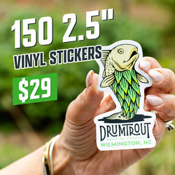 Hand holding a die-cut sticker of a stylized trout with green leafy hop scale design and text DrumTrout Brewing Company Wilmington, NC. Natural blurred greenery behind. On-image text reads 150 2.5 inch vinyl stickers for $29. High-intent keywords include craft brewery stickers, fish decals, fishing artwork stickers.