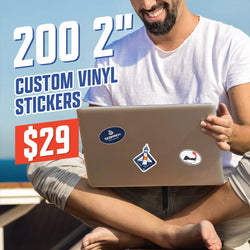 Man sitting cross-legged outdoors with bright blue ocean behind him, smiling while using a laptop decorated with small 2-inch custom vinyl stickers, including a navy oval logo, a geometric lighthouse design, and a lobster graphic. Warm sunlight highlights the scene. On-image text reads 200 2