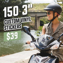Person riding a black scooter decorated with two die-cut stickers: one showing a horned skull illustration and another heart-shaped sticker with stars and crescent moon inside. Scenic river and bridge backdrop. On-image text reads 150 3 inch custom vinyl stickers for $39. Keywords: helmet stickers, scooter decals, gothic vinyl stickers, outdoor adventure stickers.