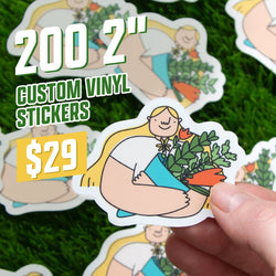 Promotional photo showing a hand holding a small sticker depicting a blonde illustrated woman hugging a bouquet of green leaves and orange flowers. Background contains multiple identical stickers scattered on green grass. White and green text reads 200 2 inch custom vinyl stickers for $29. Soft, friendly illustration style.