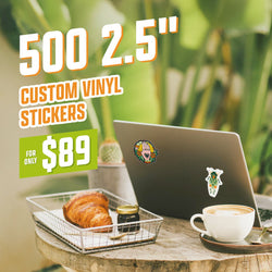 Laptop decorated with two vinyl stickers featuring colorful hand-drawn character art: one with warm oranges and expressive linework, another with a green botanical character design. Set in a cozy café scene with croissant, jam jar, and latte on a rustic table. On-image text reads 500 2.5 inch custom vinyl stickers and 89. Keywords: laptop stickers, cartoon character decals, hand-drawn vinyl art, aesthetic laptop decoration.