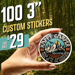 Hand holding a circular Happy Camper sticker featuring a vintage expedition badge design with distressed textures, a stylized mountain range, pine trees, a soaring eagle, and warm sunset colors. Text reads Happy Camper, Wild Life Expedition, Take Me to the Mountains, Wyoming. Forest background. On-image text reads 100 3 inch custom stickers and 29. Keywords: camping badge sticker, retro outdoor decal, mountain adventure vinyl sticker.