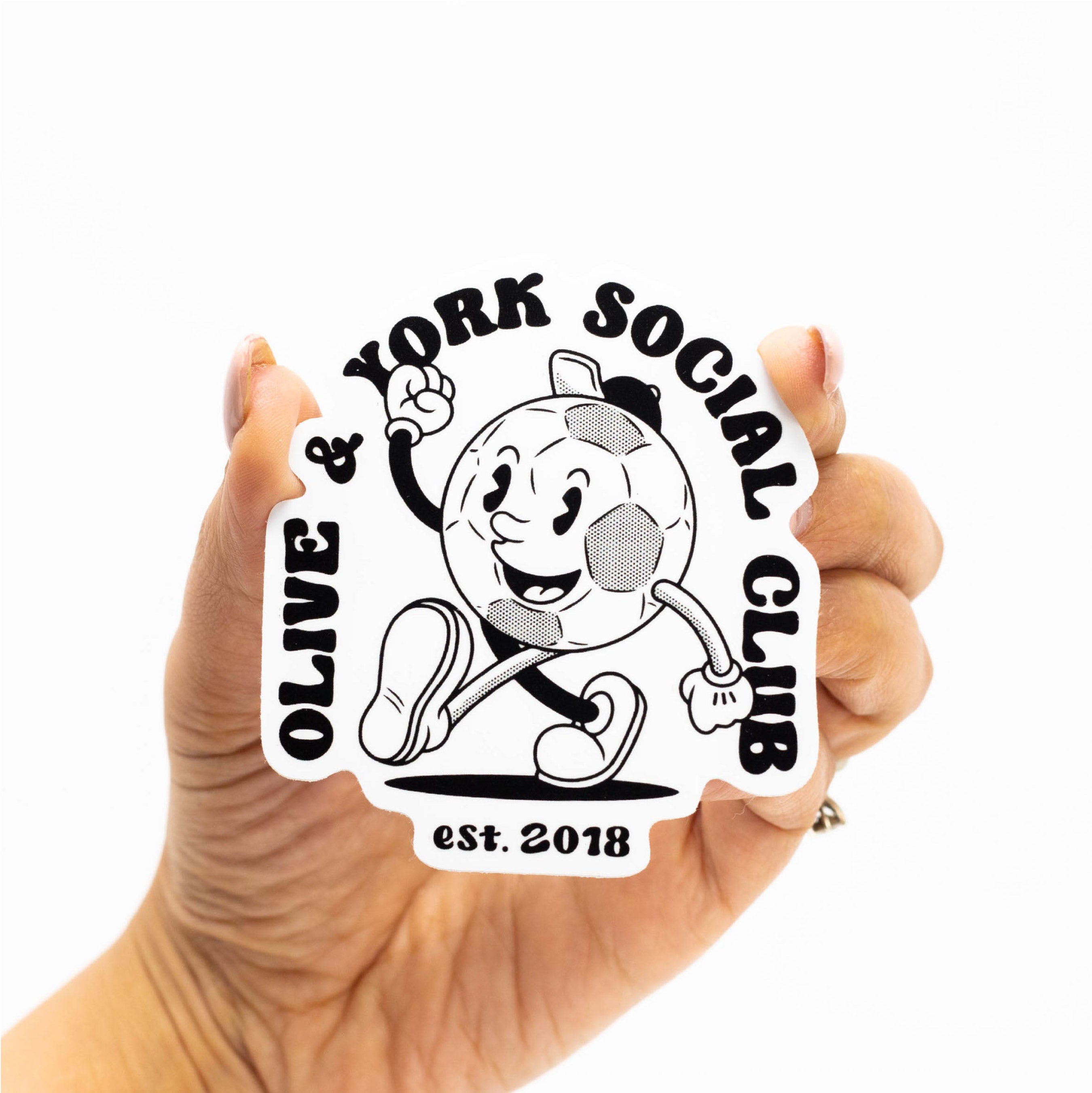 A person holds a black-and-white custom die-cut sticker featuring a smiling soccer mascot and social club branding. The sticker demonstrates custom shape cutting and sharp print quality.