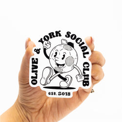 A person holds a black-and-white custom die-cut sticker featuring a smiling soccer mascot and social club branding. The sticker demonstrates custom shape cutting and sharp print quality.