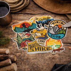 A set of colorful outdoor-themed die-cut stickers arranged on a rustic wooden surface with camping props like a metal cup, rope, hat, and logs. The stickers feature illustrated mountain scenes, a campfire, a hiking boot with the words 'TAKE A HIKE,' a mountain graphic with 'HIKEING CLUB,' and other nature motifs.