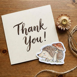 A small die-cut sticker of a cute hedgehog under a mushroom, placed on a cream-colored card that says 'Thank You!' on a rustic wooden background with twine and a dried flower.