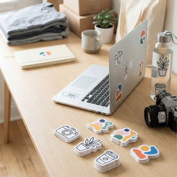 A styled desk scene with a silver laptop, camera, water bottle, folded clothing, notebook, and small plants, plus several colorful die-cut sticker shapes placed in front of the desk.