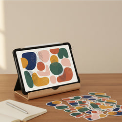 A tablet in a black case stands on a wooden desk in a warm indoor setting, displaying an abstract multicolor sticker design. Several matching stickers are scattered on the desk in front, with an open notebook and pen visible in the lower left.