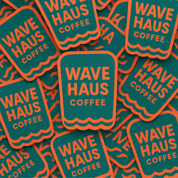 A pile of Wave Haus Coffee branded stickers or labels with a teal background, orange border, and orange text reading 'WAVE HAUS COFFEE'.