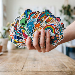 A hand holds a large, colorful fan of die-cut stickers featuring whimsical cartoon designs like a rainbow, alien, waves, and a rocket ship, set against a blurred indoor background with a wooden table.