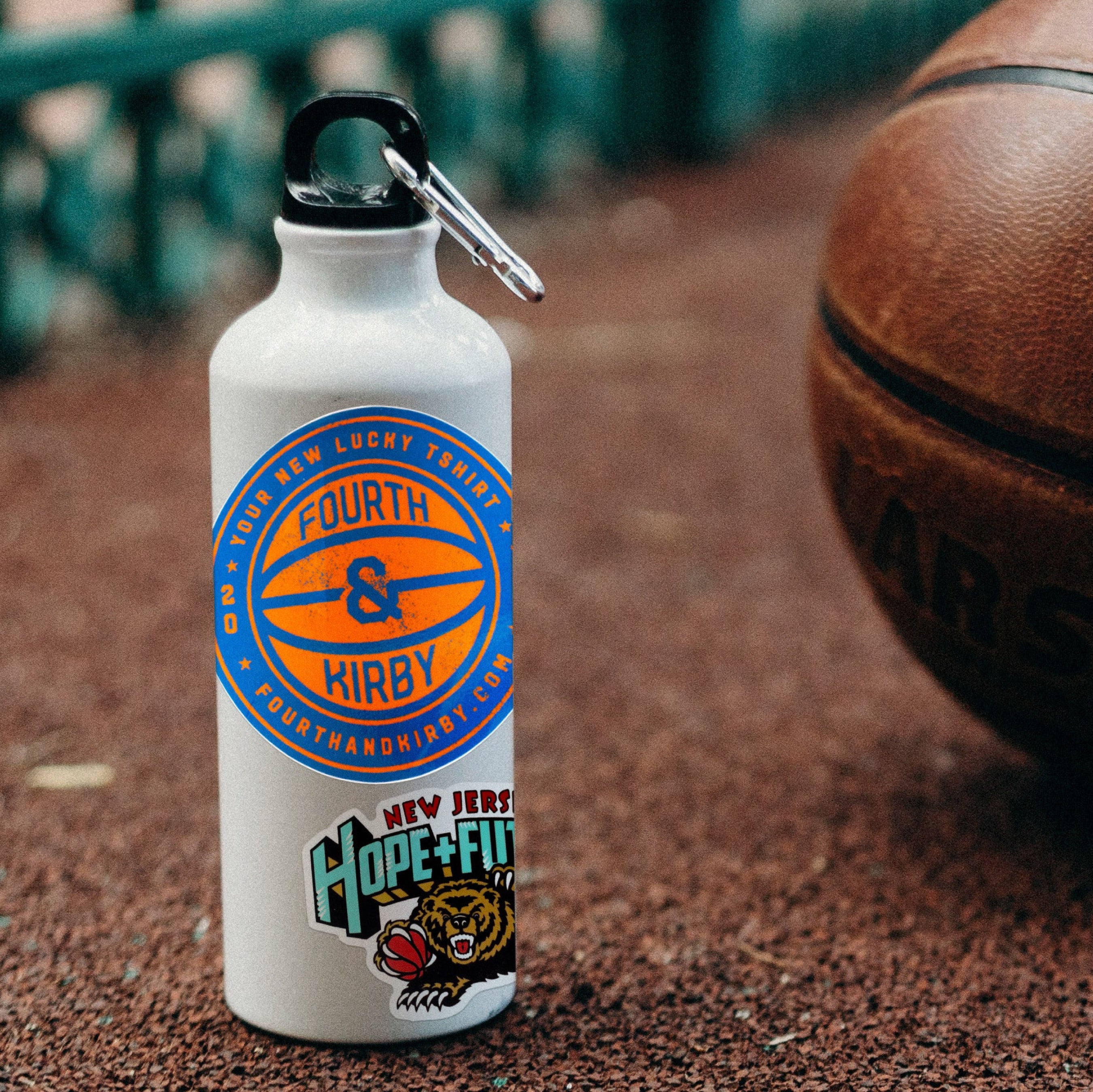 A white water bottle with a black carabiner clip, featuring custom blue and orange circular graphics and a 'New Jersey Hope & Future' bear mascot sticker, set on a brown textured ground with a basketball blurred in the background.