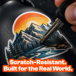 A key is dragged across a custom vinyl sticker applied to a black bottle to demonstrate scratch resistance. The colorful mountain artwork remains protected. Promotional text emphasizes real-world durability.