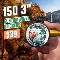 Text reads 150 3 inch custom vinyl stickers, 39 dollars. A hand holds a round embroidered-style sticker reading More Parks Less Parking with an illustration of a bear beside a lake under pine trees. Nature and conservation aesthetic, custom vinyl sticker.