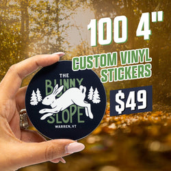 Promotional image featuring a hand holding a round dark green sticker with a white illustrated rabbit jumping between pine trees and text reading The Bunny Slope Warren, VT. Background shows sunlit forest with warm golden tones. White and green text at top states 100 4 inch custom vinyl stickers for $49.