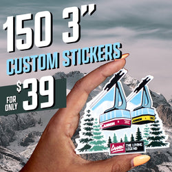 Promotional image advertising ‘150 3-inch custom stickers for only $39.’ A hand holds a die-cut sticker featuring two aerial tram cars labeled ‘Cannon 1’ (yellow) and ‘Cannon 2’ (red) ascending above snow-covered evergreen trees. At the bottom of the sticker is a logo reading ‘Cannon — The Living Legend.’ The background shows a snowy mountain landscape