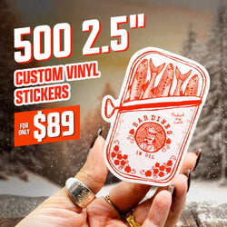 Hand holding a red and white custom vinyl sticker shaped like a sardine can with illustrated fish and floral details, reading Sardines in Oil. Promotional text on the image reads 500 2.5-inch custom vinyl stickers for only $89. Background shows an outdoor winter scene with falling snow and blurred trees.