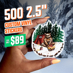 Hand holding a 2.5-inch custom vinyl sticker featuring an illustrated bear sitting by a campfire beside a red tent labeled OGE, surrounded by dark green pine trees and a snowy forest backdrop. Warm campfire colors contrast with the icy landscape. On-image text reads 500 2.5