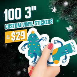 Hand with red nail polish holding two festive custom vinyl stickers, including a teal Christmas-tree character with yellow and pink ornaments and a smiling teal-and-orange holiday stocking. Background features dark green pine branches and silver star decorations. On-image text reads 100 3