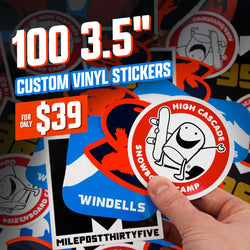 Hand displaying a round red-white-blue High Cascade Snowboard Camp sticker showing a smiling character holding a snowboard. Background shows additional Windells and MilepostThirtyFive snowboarding stickers. On-image text 100 3.5 inch custom vinyl stickers and 39. Keywords: snowboarding stickers, winter sports decals, camp vinyl sticker pack.