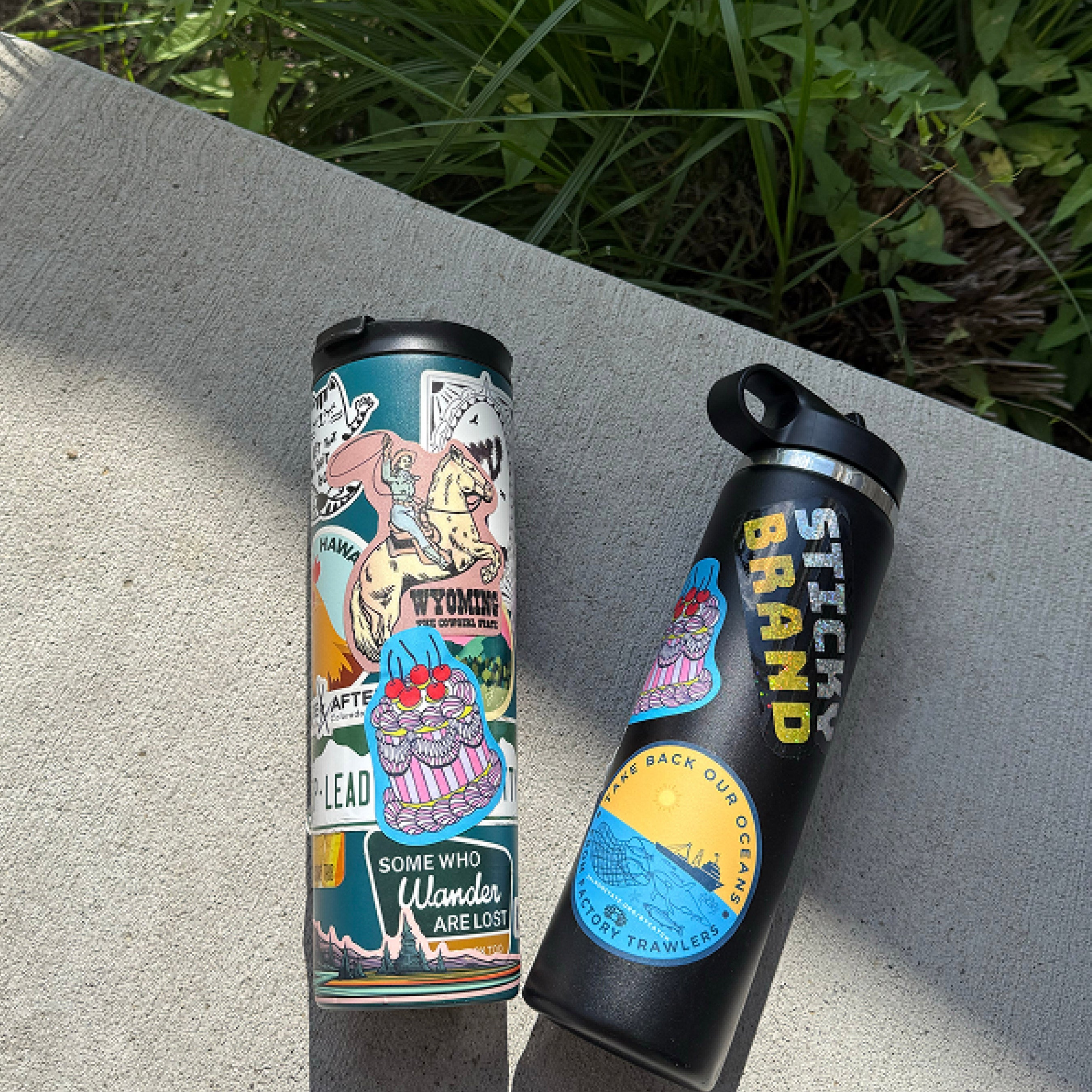 Insulated travel tumbler and reusable water bottle covered with colorful custom die-cut vinyl stickers featuring travel, outdoor, and lifestyle artwork. Visible sticker text includes "WYOMING COWBOY STATE," "HAWAII," "SOME WHO WANDER ARE LOST," "STICKY BRAND," and "TAKE BACK OUR OCEANS – VICTORY TRAWLERS." The waterproof stickers demonstrate vibrant full-color printing for decorating drinkware, water bottles, tumblers, laptops, and personal gear.