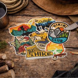 Multiple hiking and camping-themed stickers are displayed on a rustic wooden table among outdoor gear. The designs feature mountains, campfires, hiking boots, tents, and wilderness artwork.