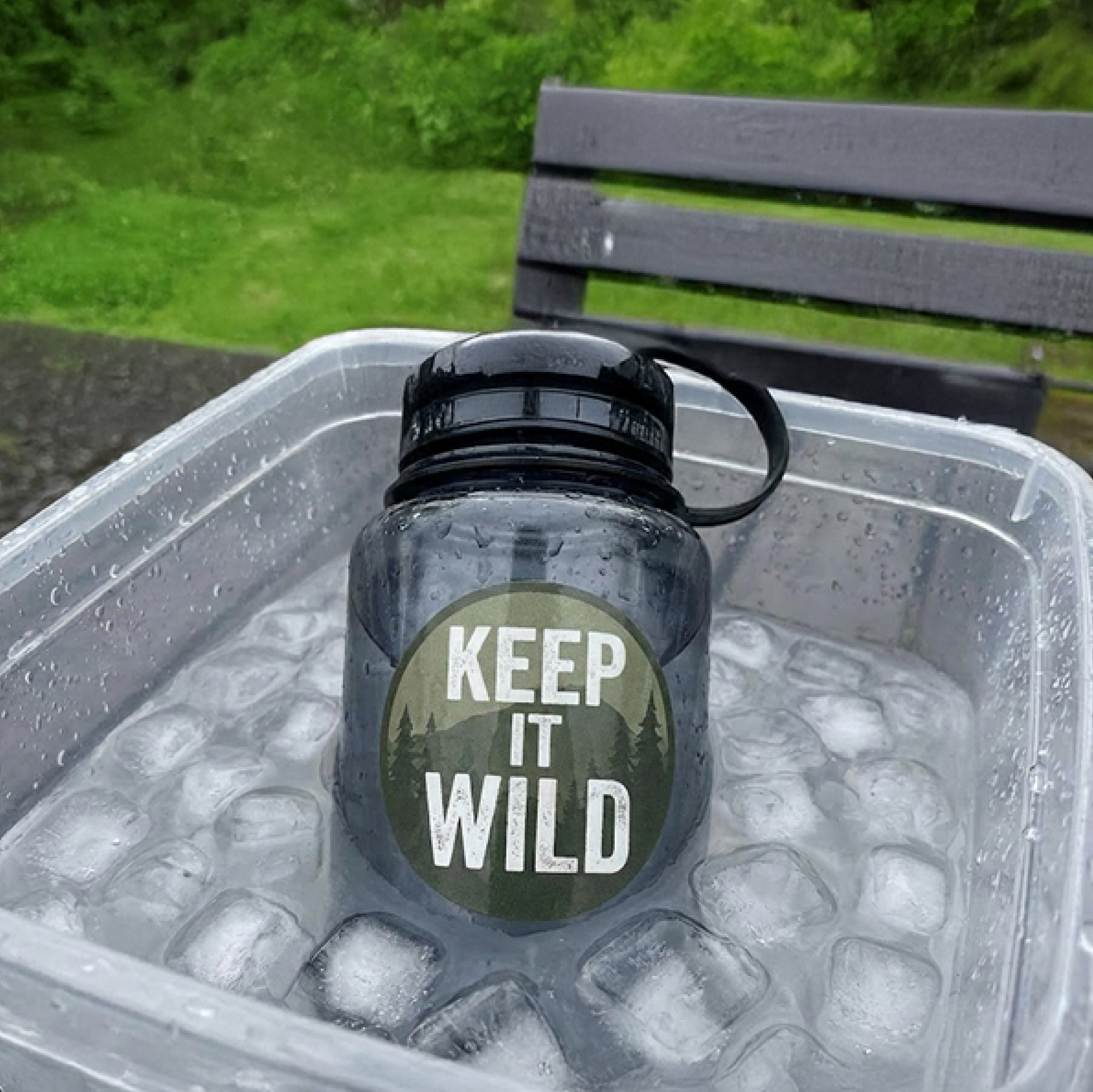 A reusable water bottle sits partially submerged in a clear container filled with ice and water outdoors. A circular sticker on the bottle reads "Keep It Wild" and features a forest-themed design. Water droplets cover the bottle while greenery and a park bench appear in the background. The image demonstrates the waterproof and weather-resistant performance of custom vinyl stickers in cold and wet conditions.