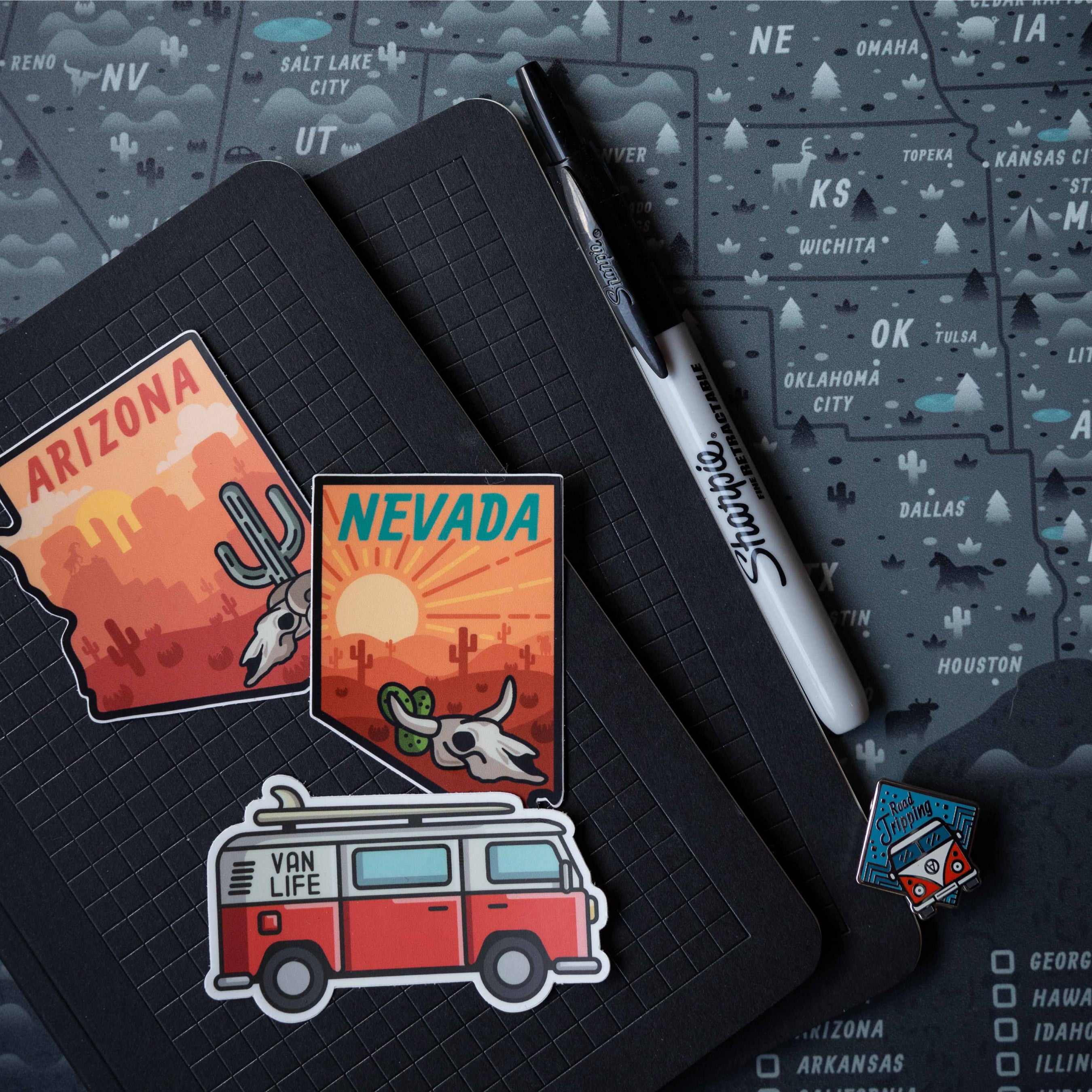 A notebook covered with colorful travel and state-themed stickers rests on a map. The stickers feature desert landscapes, van life artwork, and adventure-inspired illustrations.