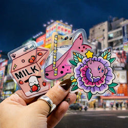 A hand holding three colorful kawaii-style clear vinyl stickers in front of a nighttime city street with bright neon signs. The stickers include a pink milk carton labeled Milk with strawberries and a small character, a pink drink in a glass with a striped straw, and a cute purple character surrounded by flowers and leaves.