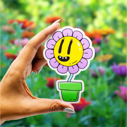 A hand holds a colorful die-cut sticker featuring a smiling yellow flower with purple petals in a green flowerpot. A vibrant, softly blurred garden filled with colorful flowers appears in the background.