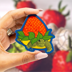 A hand with light pink manicured nails holds a die-cut sticker shaped like a green turtle crawling inside a large red strawberry with green leaves, outlined in blue. The background is a soft-focus, blurred image of more strawberries.