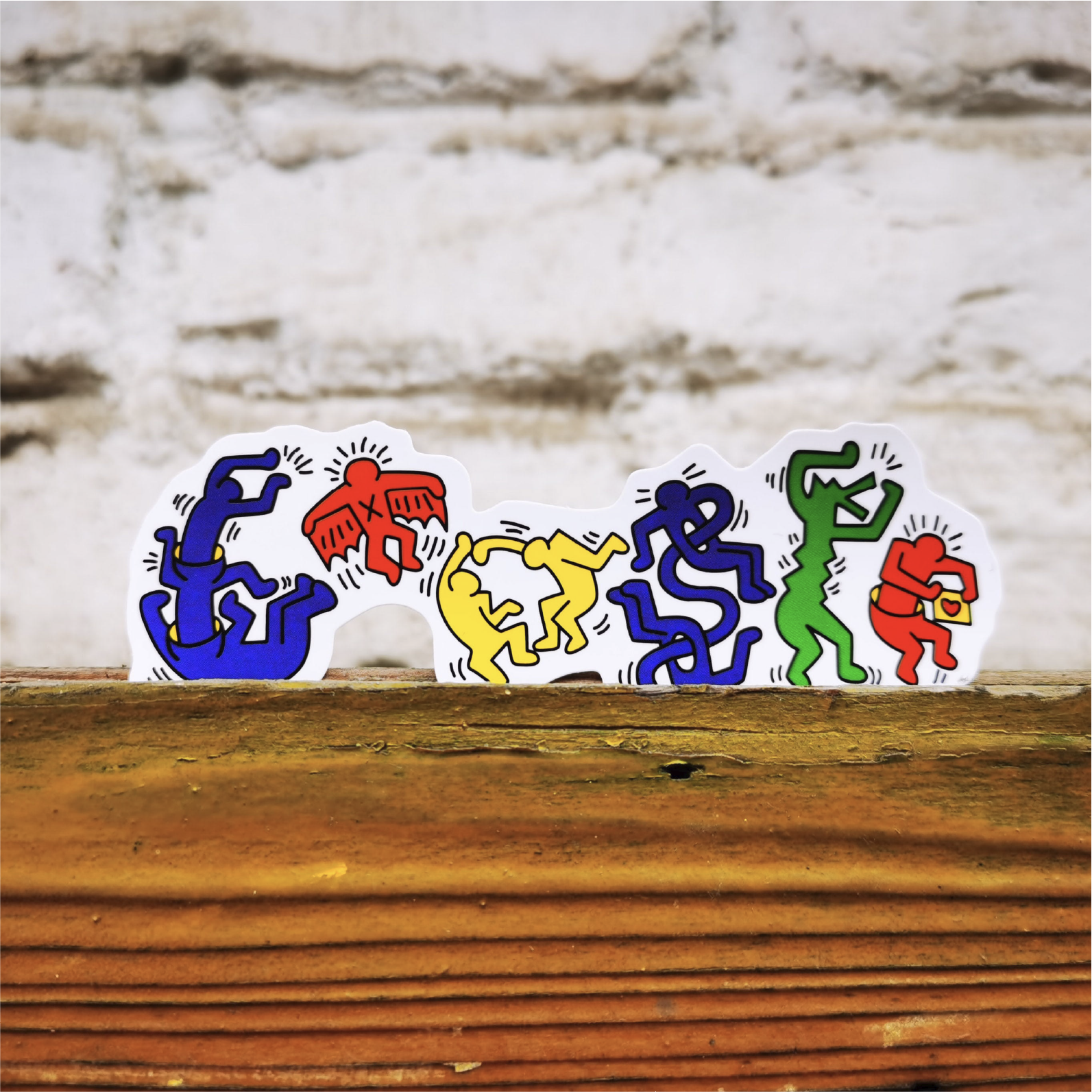 A row of five Keith Haring style stickers in blue, red, yellow, green, and orange, displayed on a rustic wooden surface against a white brick wall background.