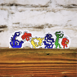 A row of five Keith Haring style stickers in blue, red, yellow, green, and orange, displayed on a rustic wooden surface against a white brick wall background.
