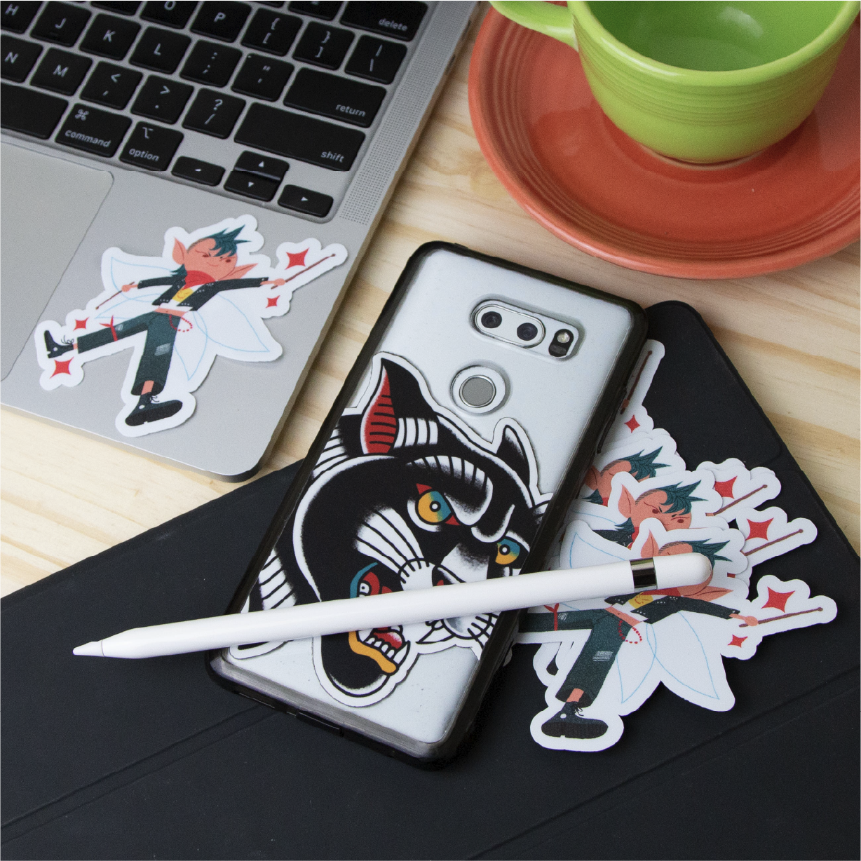Custom die-cut stickers displayed on everyday personal items. A laptop features a colorful character sticker, while a smartphone has a large illustrated black cat sticker. Additional character stickers rest on a black tablet beside a white stylus, with a green cup and saucer nearby.