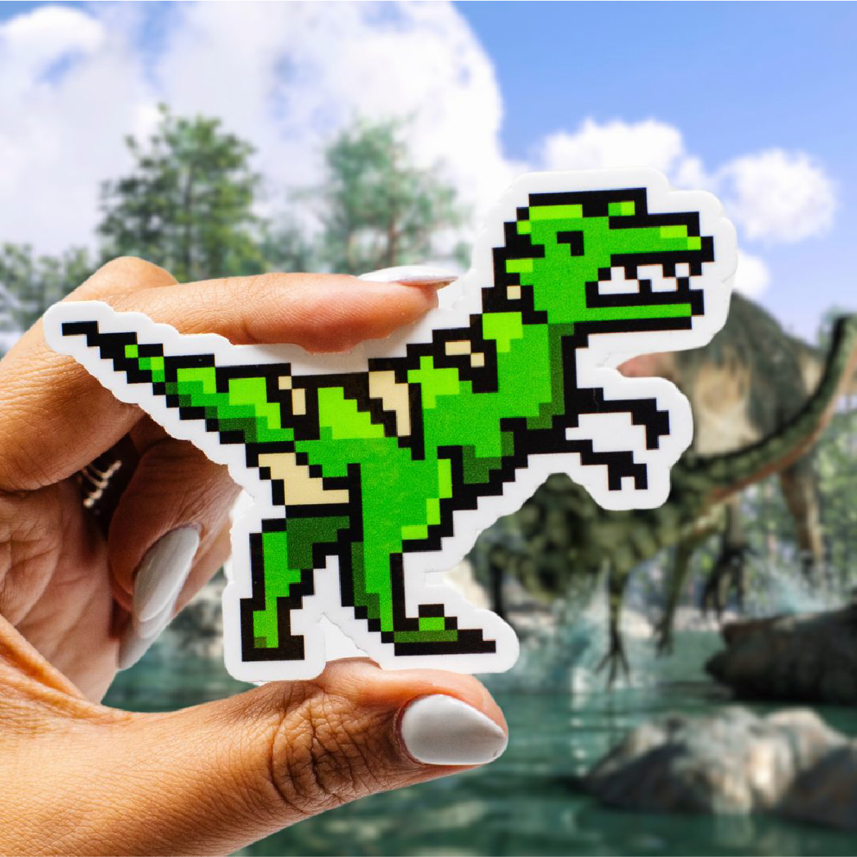 A hand holds a die-cut sticker featuring a green pixel-art dinosaur with black outlines and a white border. A prehistoric landscape with large dinosaurs, water, rocks, trees, and a blue sky appears softly blurred in the background.