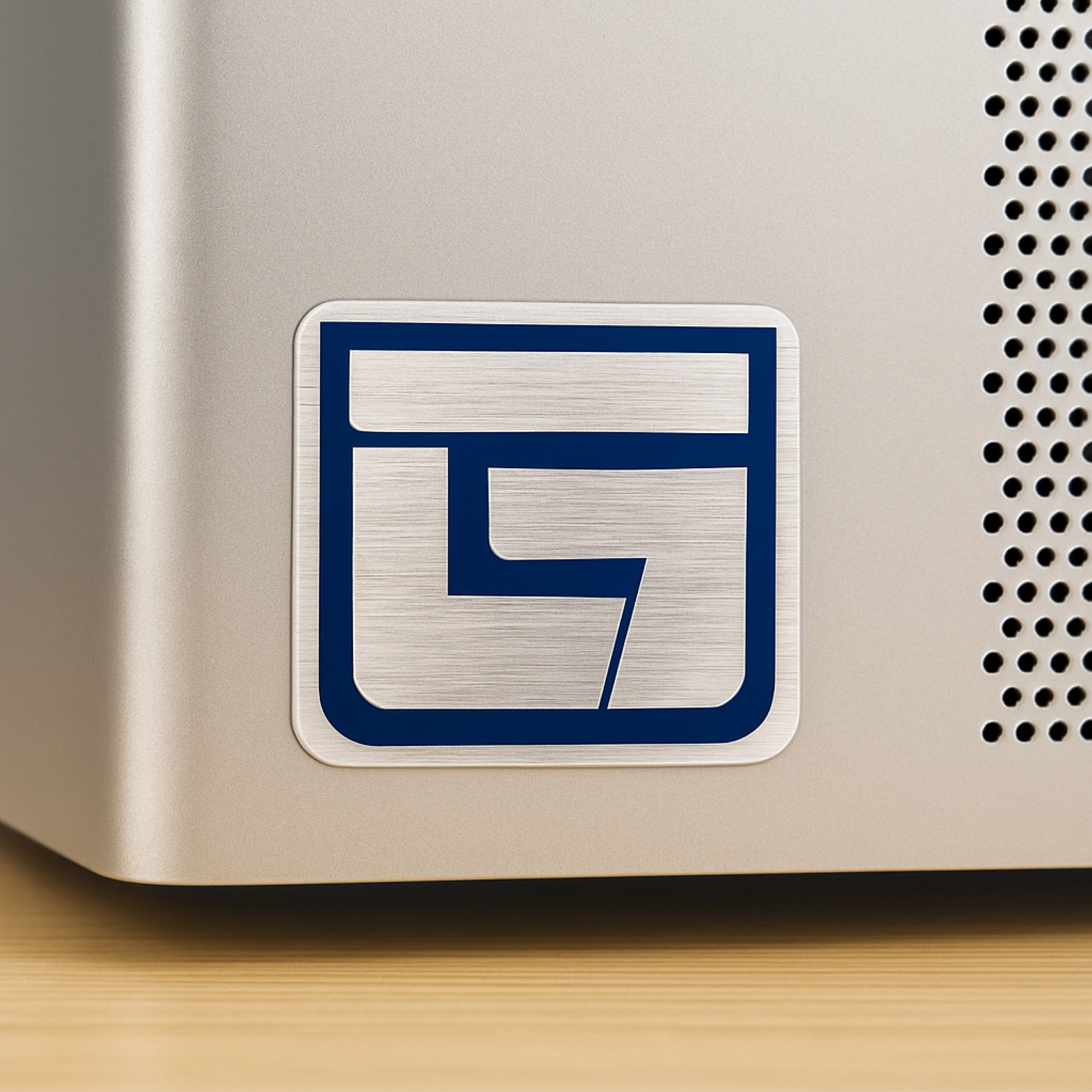 Custom brushed aluminum sticker applied to a metallic electronic device, featuring a bold blue geometric logo. The brushed silver finish blends seamlessly with the equipment surface while maintaining crisp edges and strong contrast, demonstrating professional branding and durable metal-look labeling.
