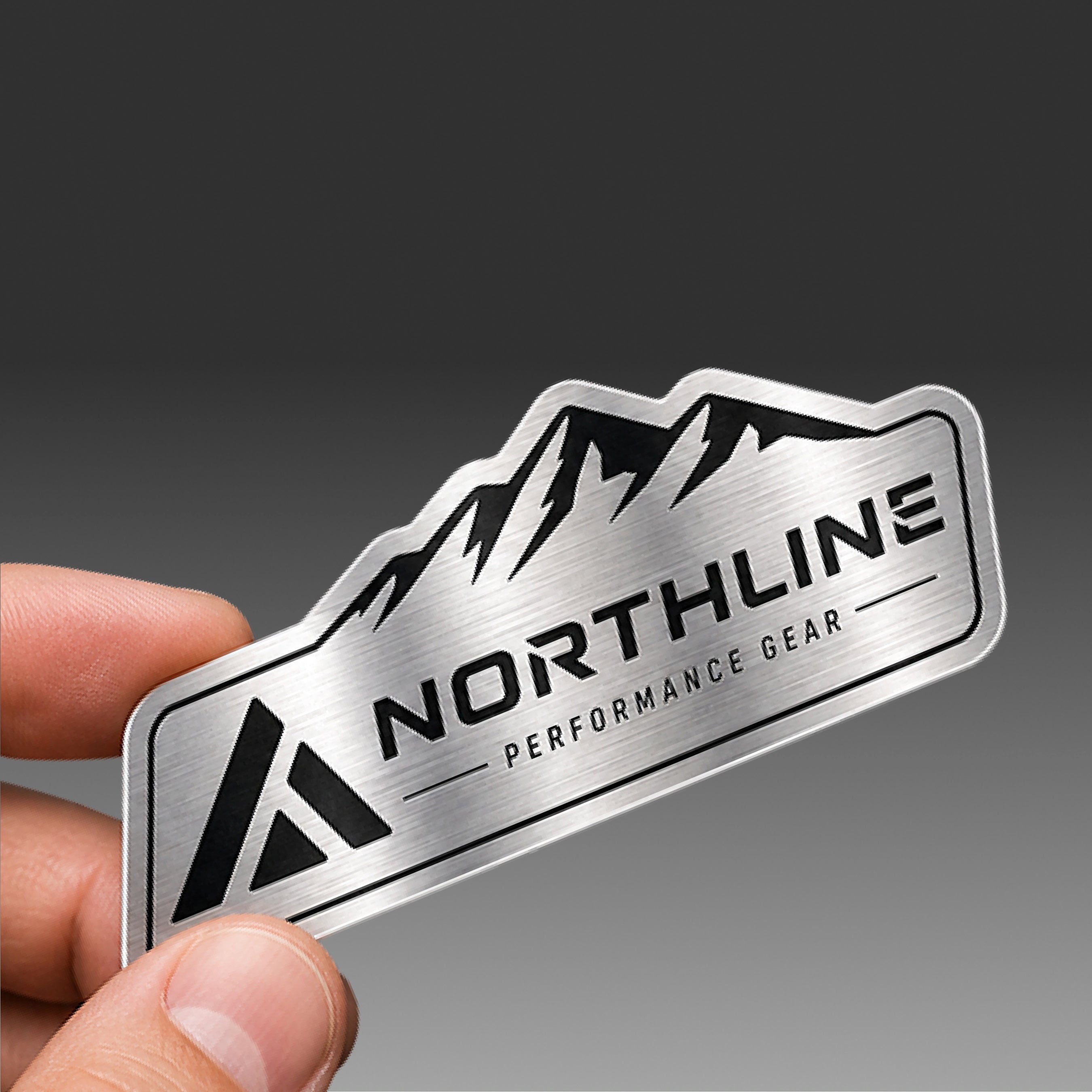 Hand holding a custom brushed aluminum sticker with a metallic silver finish, mountain graphic, and readable text that says Northline Performance Gear. The brushed metal texture creates a premium industrial look against a smooth gray gradient background, highlighting the reflective aluminum material and custom die cut shape.