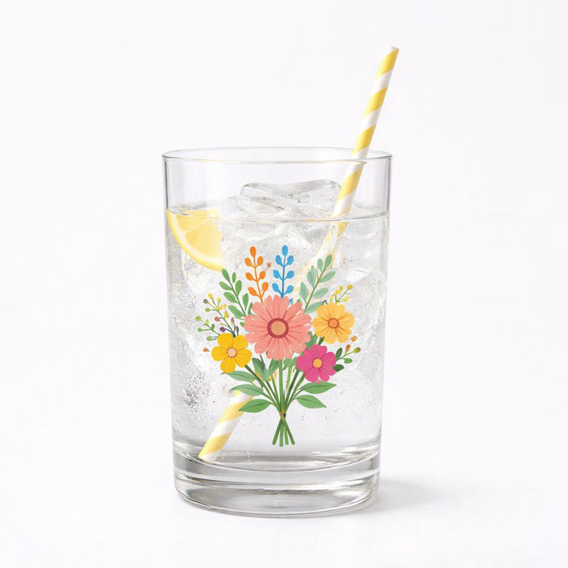 A drinking glass filled with ice and a lemon slice, featuring a transparent floral sticker on the glass. The sticker shows a bouquet of illustrated flowers in pink, yellow, orange, and blue with green leaves. A yellow-and-white striped paper straw is in the glass