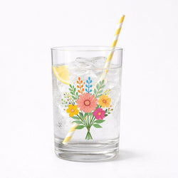 A drinking glass filled with ice and a lemon slice, featuring a transparent floral sticker on the glass. The sticker shows a bouquet of illustrated flowers in pink, yellow, orange, and blue with green leaves. A yellow-and-white striped paper straw is in the glass