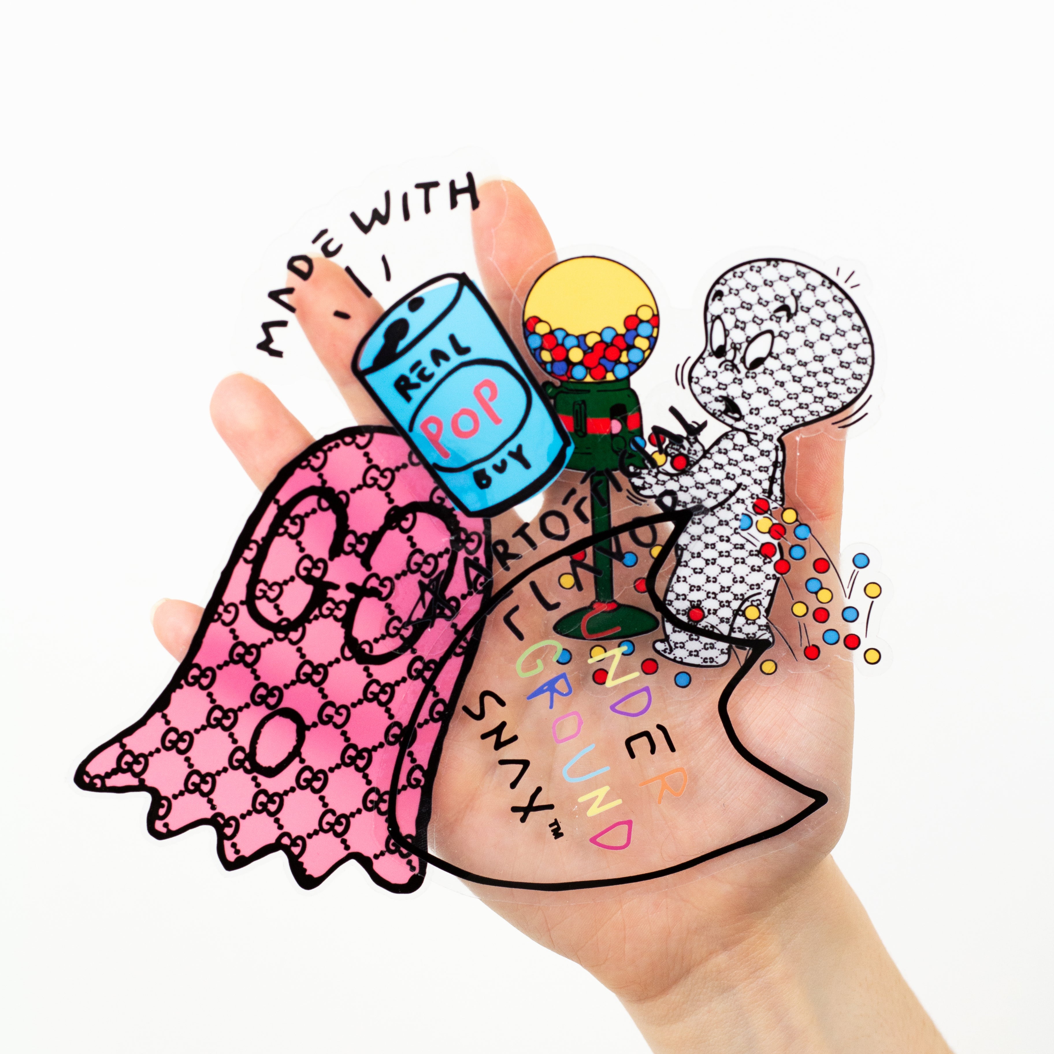 A hand holding several overlapping clear vinyl stickers against a white background. The stickers feature playful illustrated designs including a pink patterned ghost, a soda can labeled Real Pop with the phrase Made with Real Pop, a gumball machine character spilling colorful gumballs, and layered text reading Support Your Local Ground Snacks in multicolored lettering.