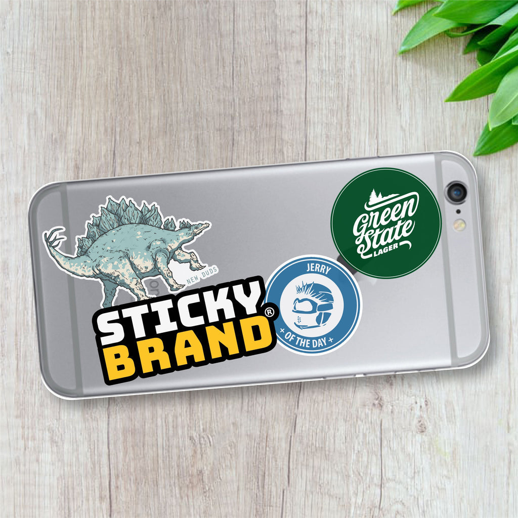 Phone case with various stickers including a dinosaur, 'Sticky Brand' logo, and 'Green State Lager' label on a wooden surface.