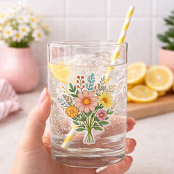 A hand holding a clear drinking glass filled with ice and a lemon slice, featuring a transparent floral sticker on the glass. The sticker shows a bouquet of illustrated flowers in pink, yellow, orange, and blue with green leaves. A yellow-and-white striped paper straw is in the glass, with a softly blurred kitchen background and lemons on a cutting board.