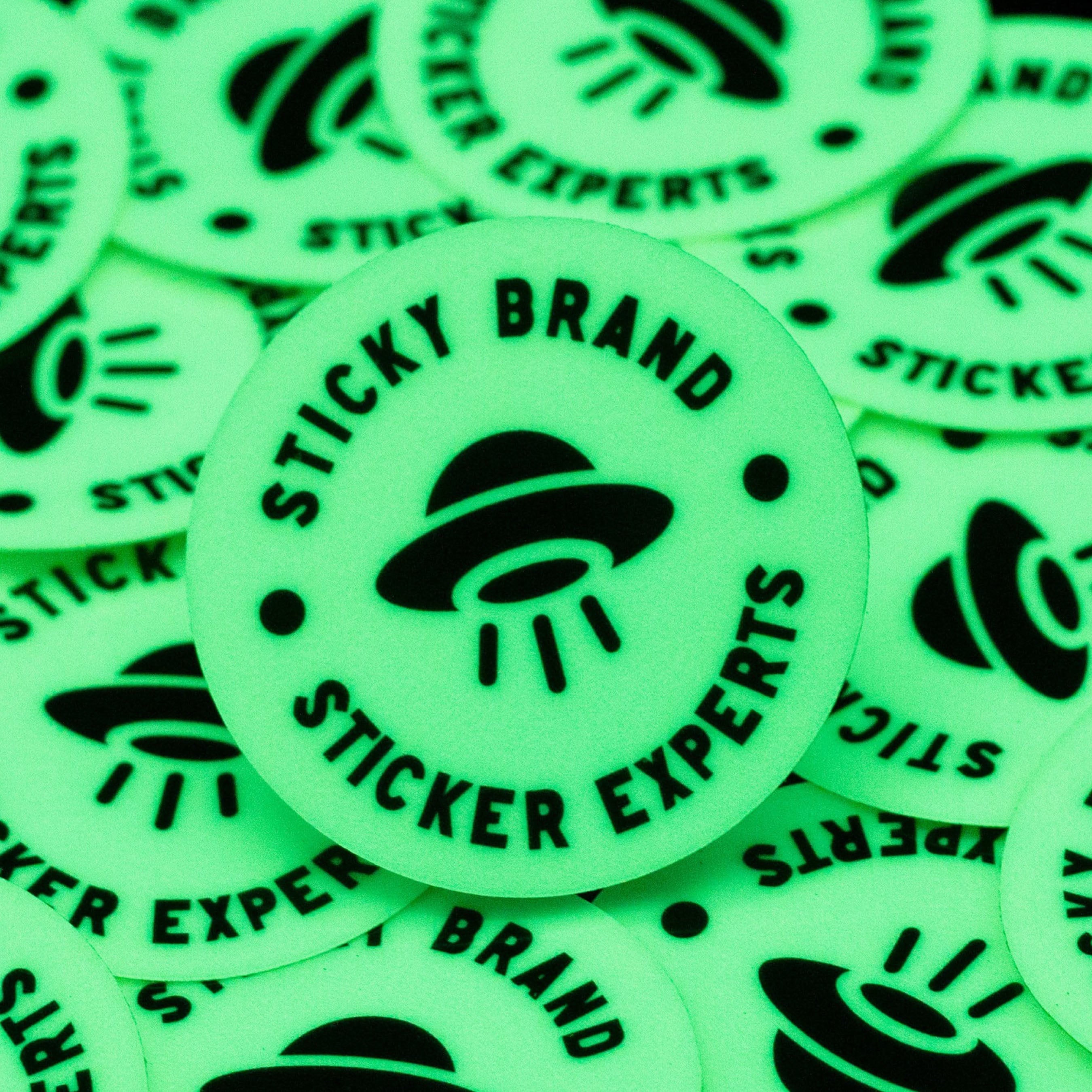 Close-up of custom glow-in-the-dark stickers featuring the Sticky Brand UFO logo illuminated in a dark environment. The photoluminescent material emits a bright green glow while maintaining sharp black graphics and crisp text. Displayed in a scattered arrangement, this image highlights the strong nighttime visibility, smooth die-cut shape, and eye-catching glow effect that makes glow-in-the-dark stickers ideal for promotions, events, product branding, and novelty applications.