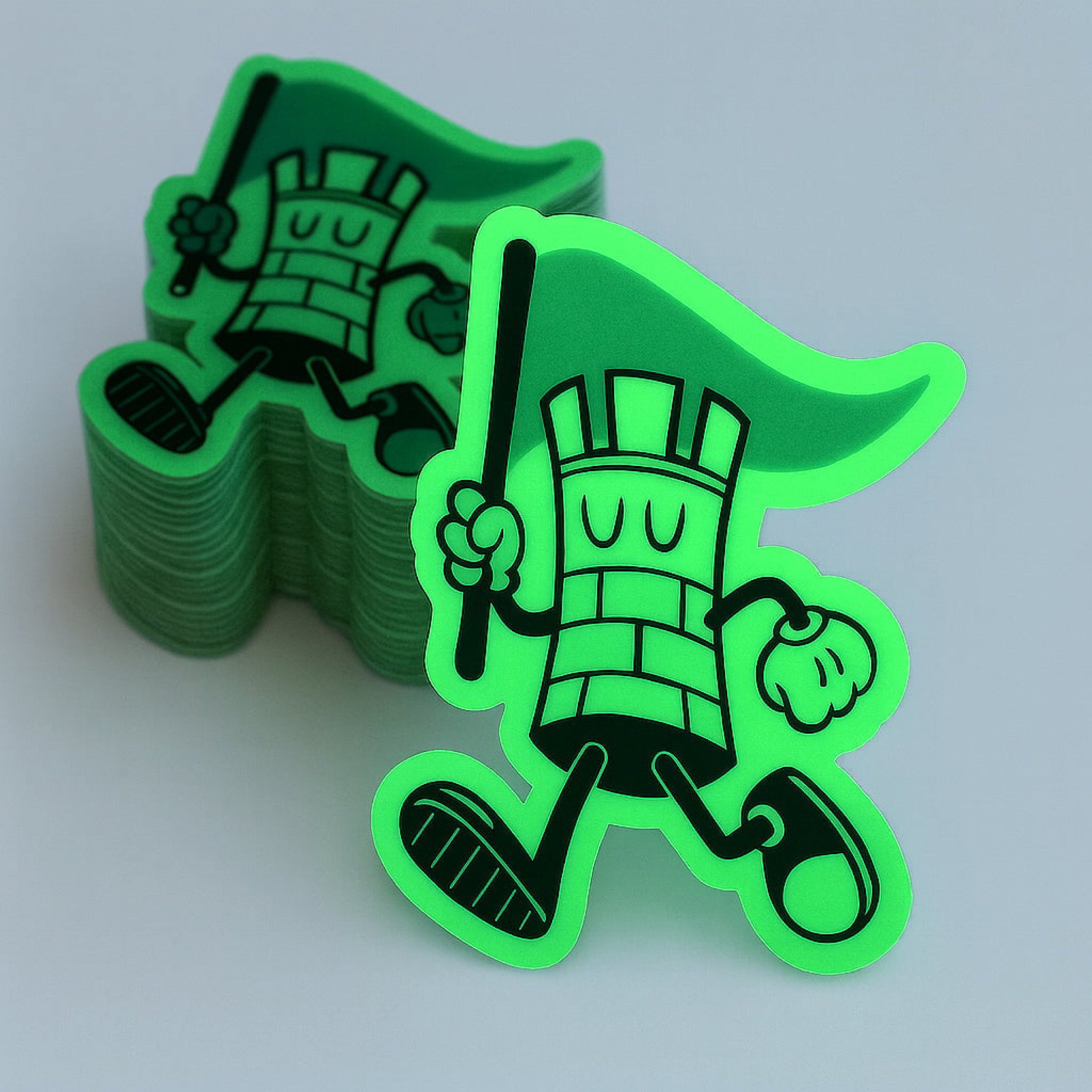 Stack of custom glow-in-the-dark die-cut stickers featuring a playful mascot character holding a flag. The specialty glow material shines brightly in low-light conditions, creating a vibrant green appearance that enhances the sticker’s outline and overall visibility. Shown alongside a stack of finished stickers, this image showcases the durability, precision cutting, and striking nighttime effect of glow-in-the-dark stickers for branding, giveaways, packaging, and promotional products.