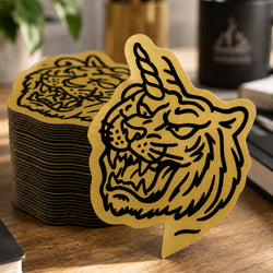 A stack of brushed gold metallic die-cut stickers features a detailed illustration of a roaring tiger with a unicorn horn in bold black artwork. The image highlights the reflective metallic finish, precision die-cutting, and premium appearance of custom brushed gold stickers.