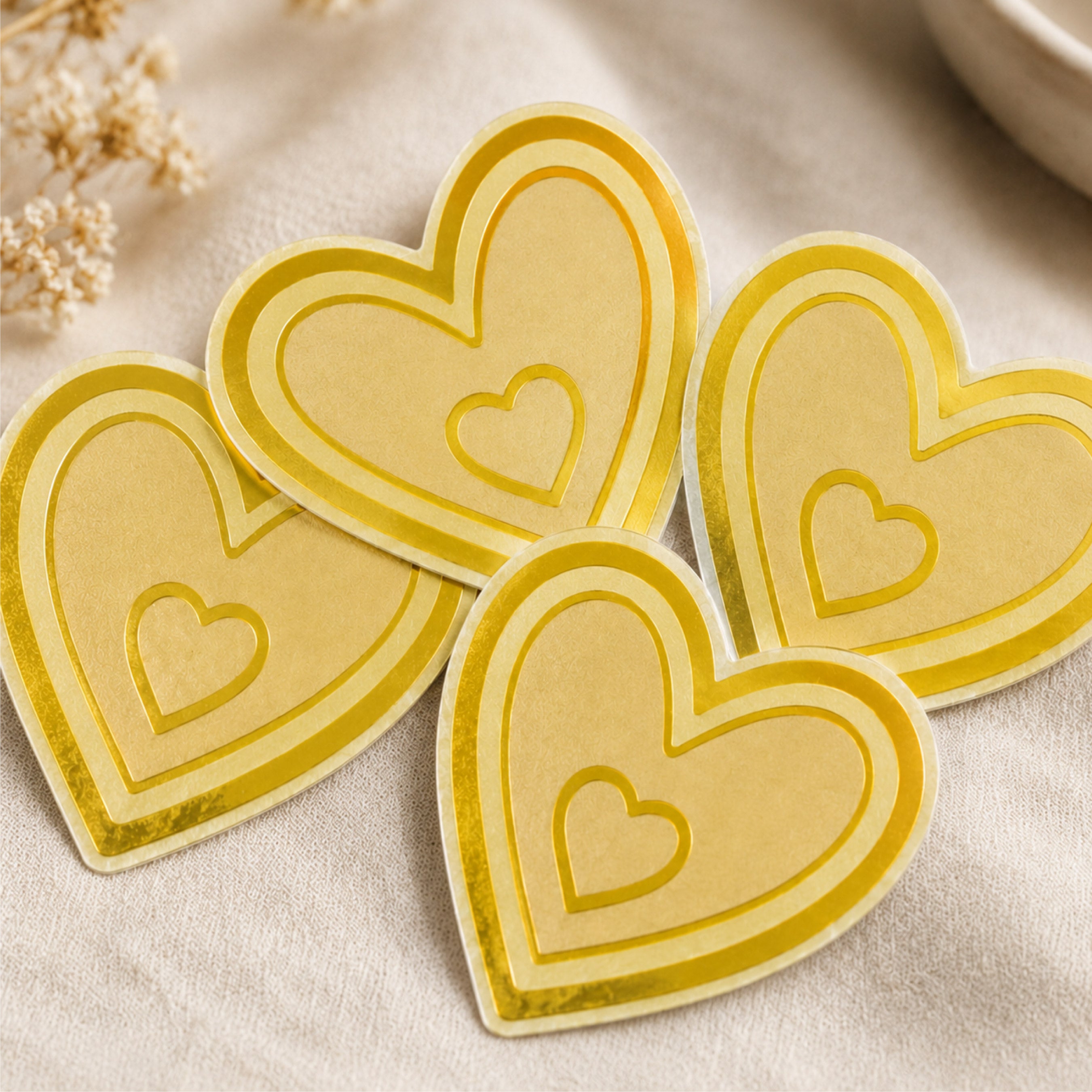 A collection of four  gold metallic heart-shaped stickers is arranged on a soft neutral fabric surface. Each die-cut sticker features a layered heart design with a smaller heart centered inside, showcasing the reflective brushed gold finish and precision-cut edges. The image highlights the premium metallic appearance and elegant look of custom brushed gold stickers for weddings, boutiques, gifts, luxury packaging, branding, and decorative applications.
