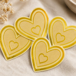 A collection of four  gold metallic heart-shaped stickers is arranged on a soft neutral fabric surface. Each die-cut sticker features a layered heart design with a smaller heart centered inside, showcasing the reflective brushed gold finish and precision-cut edges. The image highlights the premium metallic appearance and elegant look of custom brushed gold stickers for weddings, boutiques, gifts, luxury packaging, branding, and decorative applications.