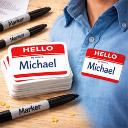 Stack of Hello My Name Is name tag stickers with the name Michael on a wooden table next to black markers, with one sticker applied to a blue shirt showing how the writable label is used for events and identification