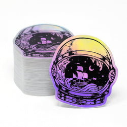 Holographic sticker of an astronaut helmet with a ship design, next to a stack of similar stickers. Rainbow iridescent sailboat waves maritime