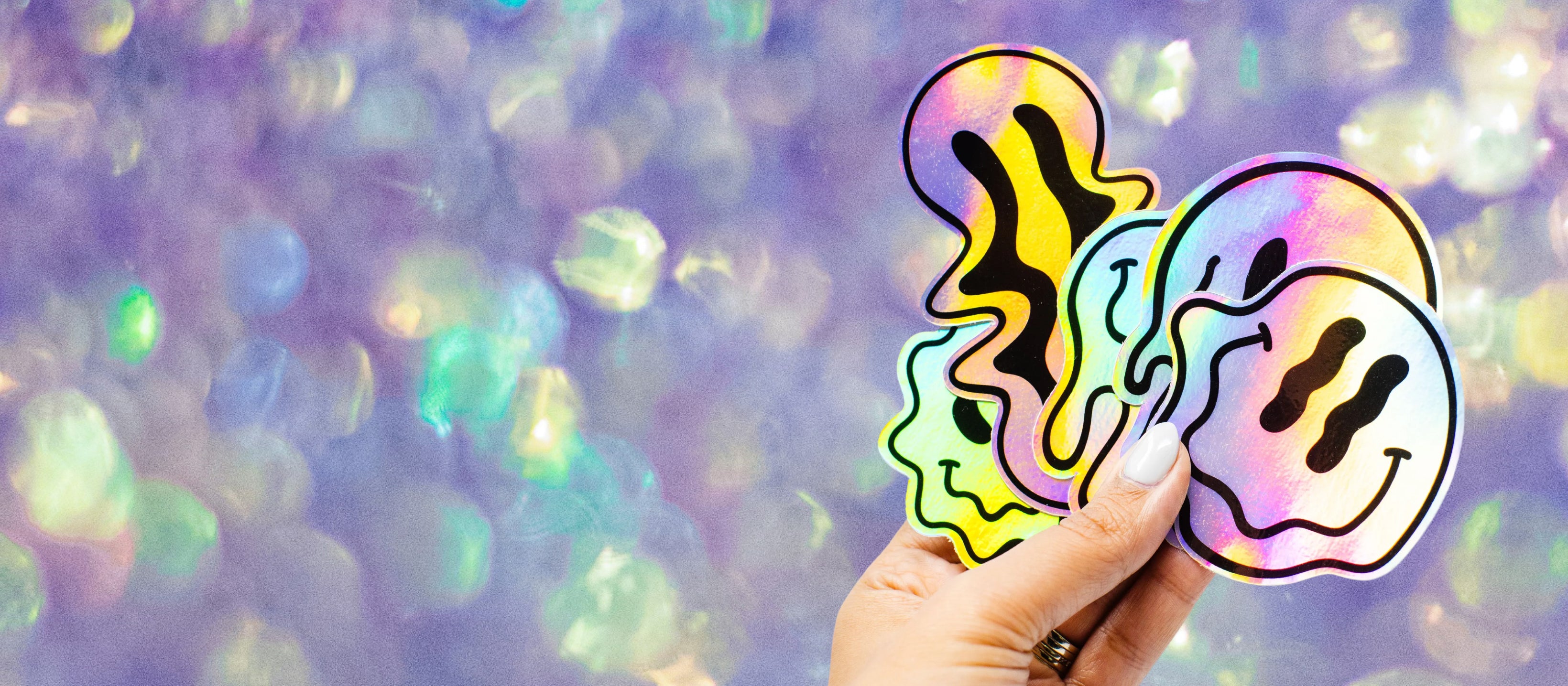 Promotional banner image featuring a hand holding five overlapping holographic smiley-face stickers with black abstract facial details. The stickers reflect rainbow colors against a soft purple and iridescent bokeh-style background. No readable text appears on the image. The scene highlights holographic vinyl material, glossy finish, bold outlines, and vibrant reflective effects used for custom die-cut stickers.